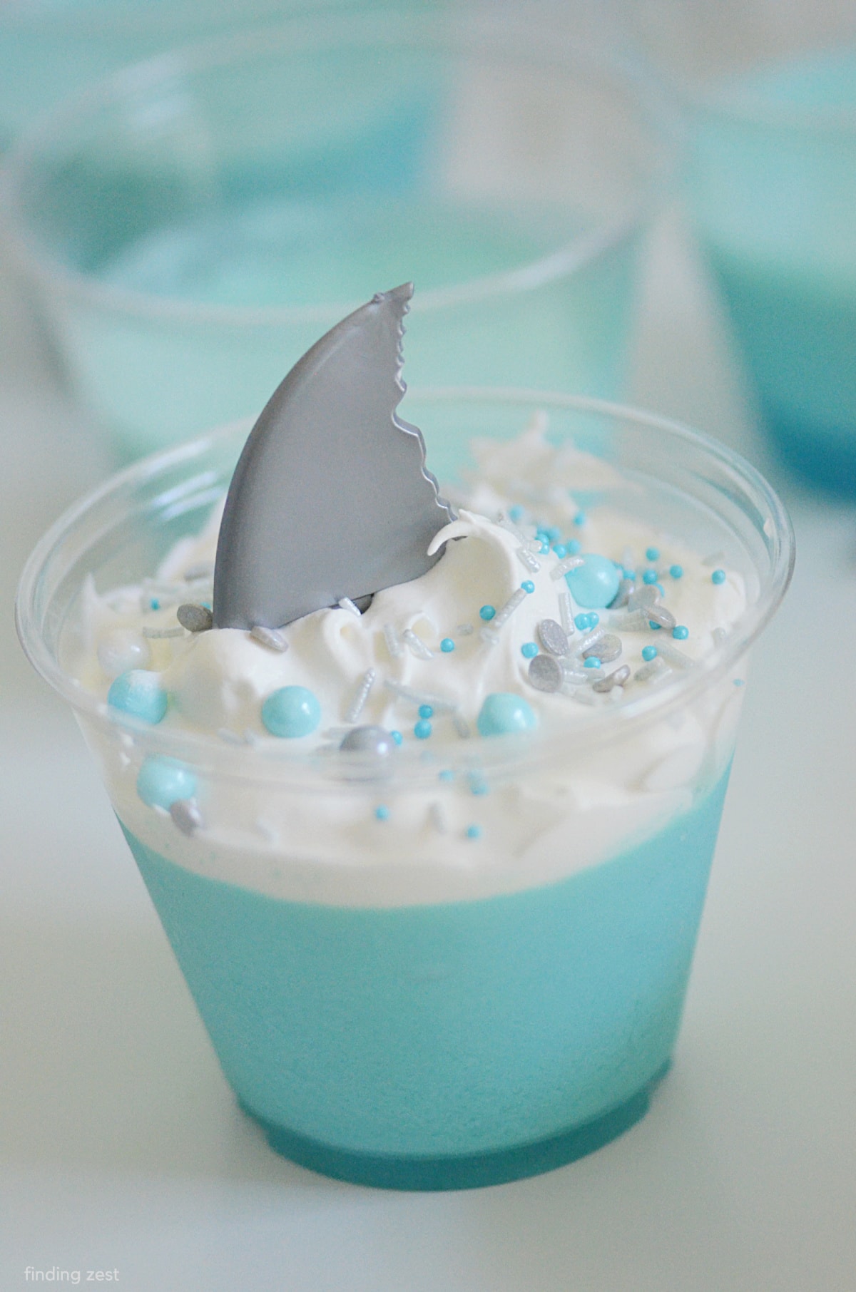 Shark Jello Cups are a simple way to create fun shark themed desserts for your guests. This blue jello mousse is super easy to whip up and looks great with whipped topping waves and colorful sprinkles. Whether you are having a traditional shark party or baby shark party theme, your party is sure to be swimming with fun!