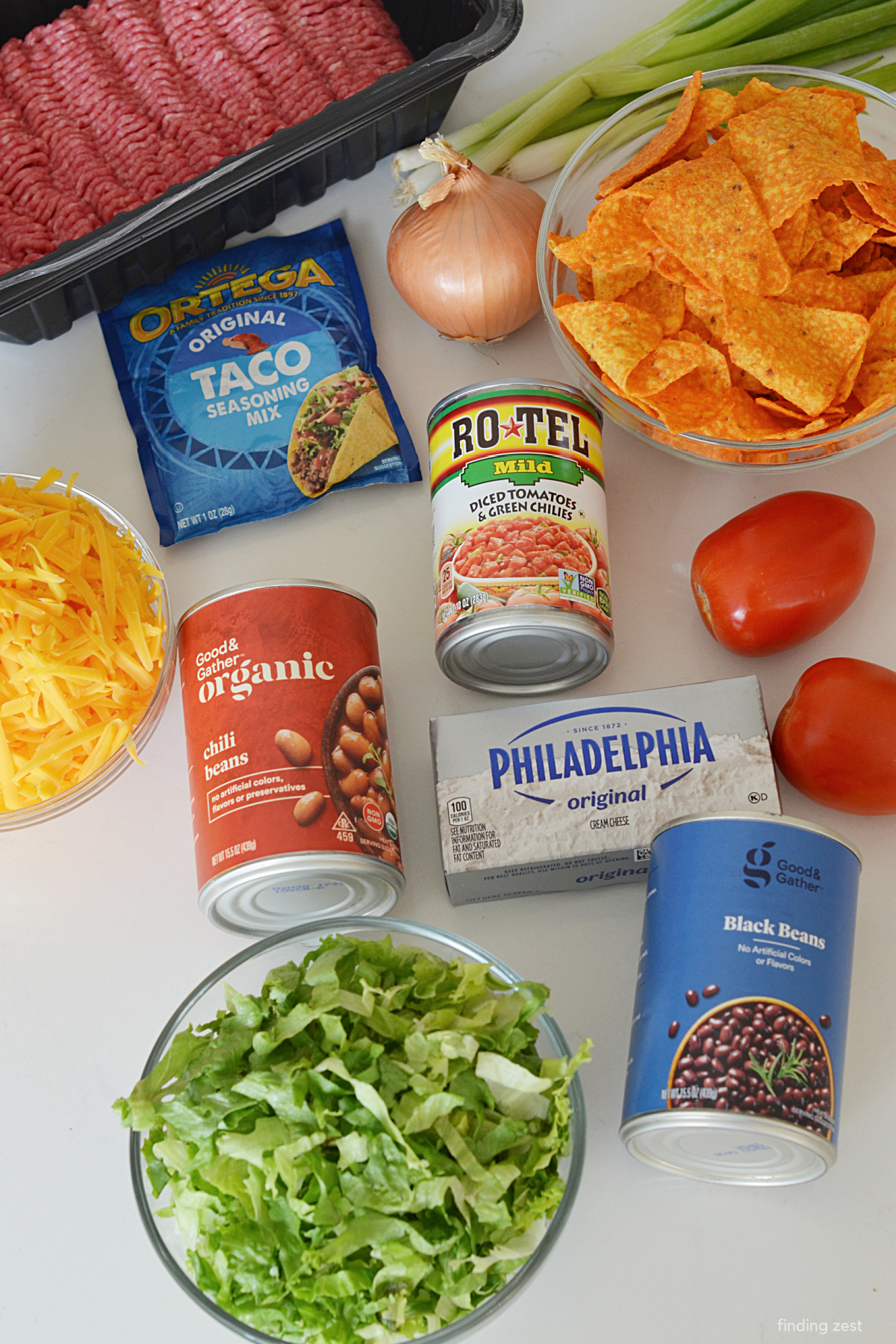 Taco casserole ingredients including ground beef, Doritos, black beans, chili beans, taco seasoning, cream cheese and fresh veggies