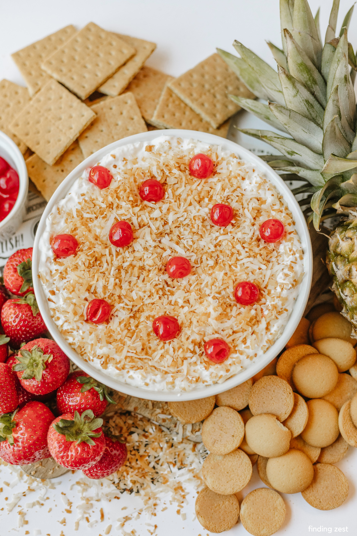 Creamy pineapple coconut dip garnished with toasted coconut and a pineapple top, surrounded by fresh strawberries, graham crackers and vanilla wafers.